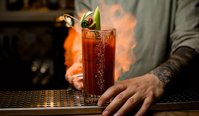 A person with a tattooed arm holds a tall glass of a red cocktail garnished with celery and herbs, with smoke rising around the drink on a bar counter.