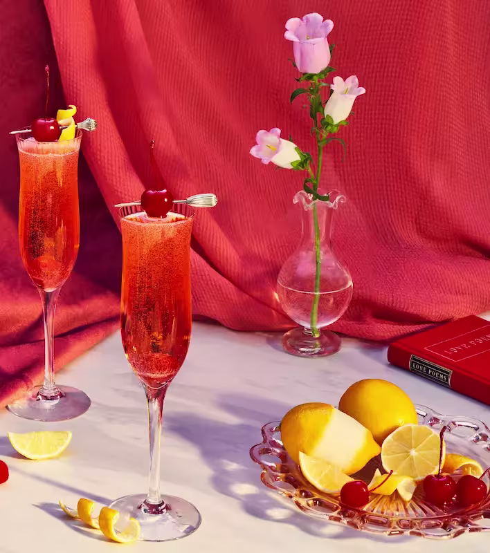 Two champagne flutes with pink cocktails and cherries are on a marble surface, alongside lemon wedges, more cherries, a glass vase with a pink flower, and a red book.