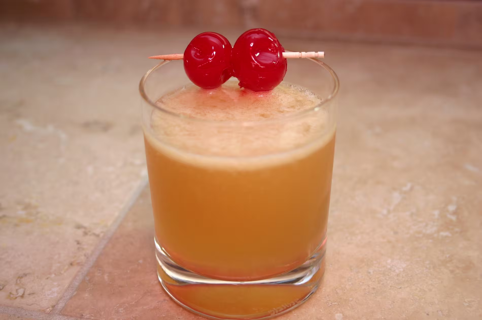 A glass of orange-brown cocktail topped with two maraschino cherries on a toothpick, set on a light-colored countertop.