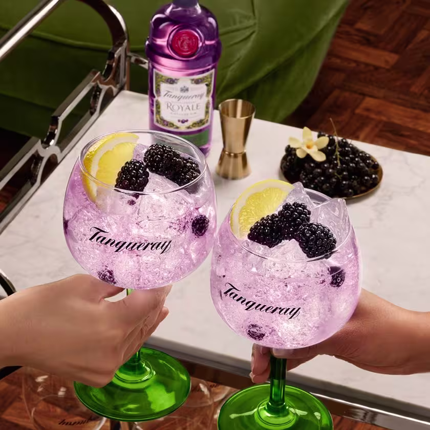 La imagen muestra un brindis con dos copas que contienen cócteles preparados con gin Tanqueray, mientras que al fondo se aprecia la botella de gin Tanqueray Royale y otros elementos de coctelería.