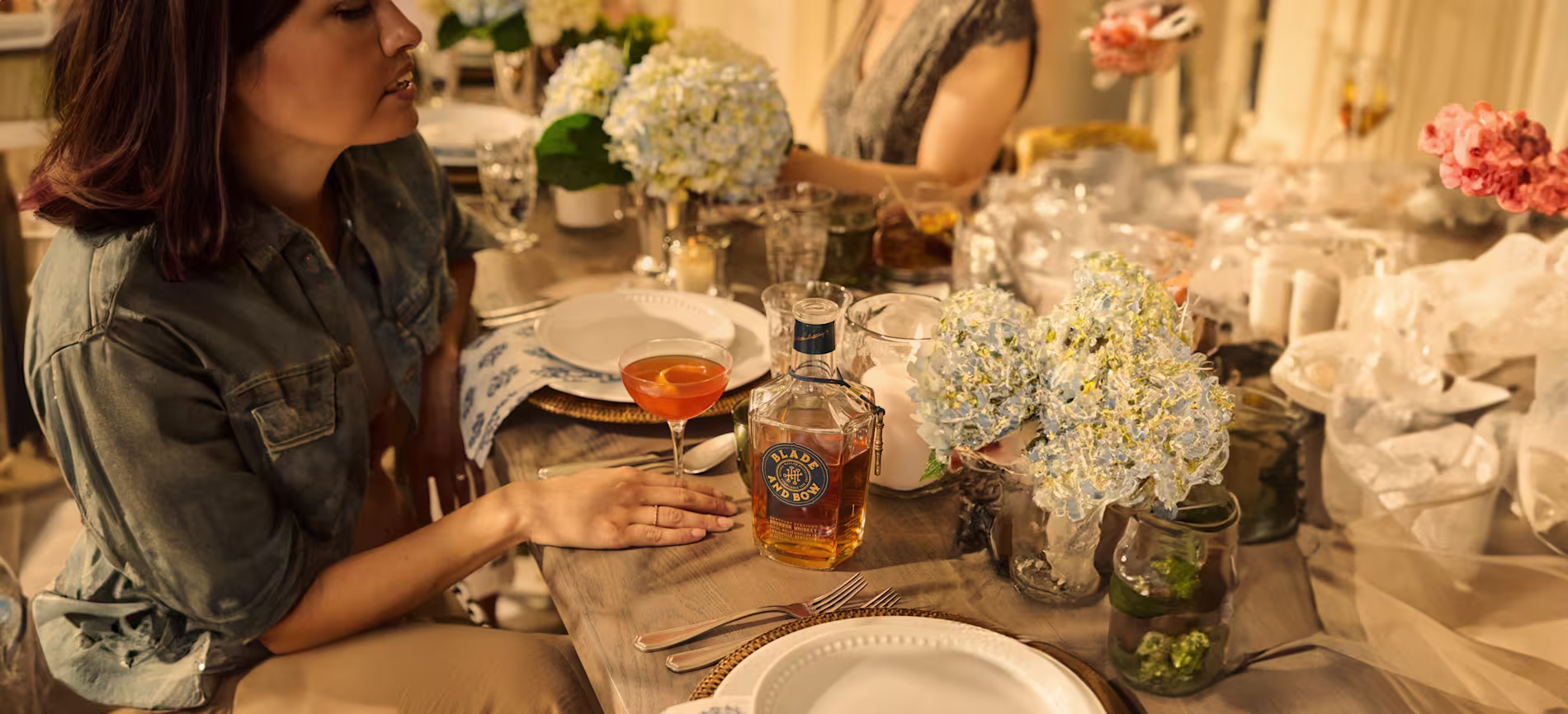 A woman sits at a decorated dining table with plates, cutlery, flowers, a cocktail, and a bottle of Royal Salute whisky.