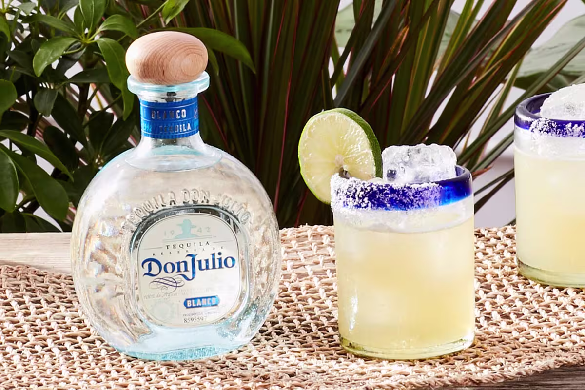 A bottle of Don Julio Blanco tequila sits next to a glass of margarita with ice and a lime wheel, on a woven mat with greenery in the background.