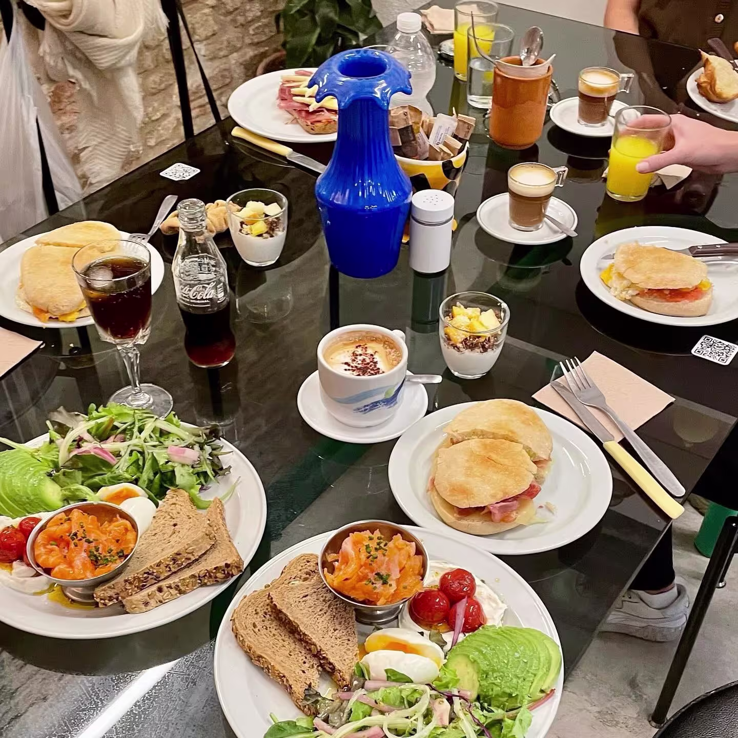 Mesa de cristal con platos variados de desayuno: tostadas con salmón, ensaladas frescas, yogur con fruta, cafés, zumos y bollería, en un ambiente acogedor y luminoso.