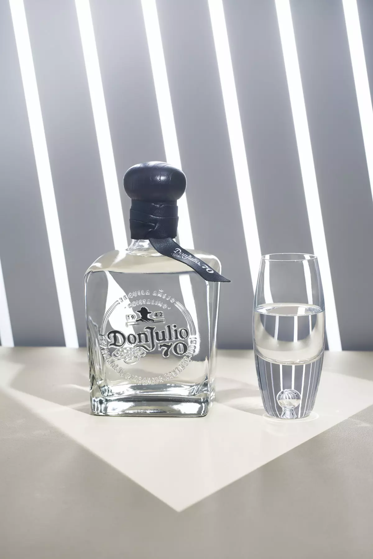 A bottle of Don Julio 70 tequila stands next to a clear glass filled with tequila on a light-colored surface with vertical white light stripes in the background.