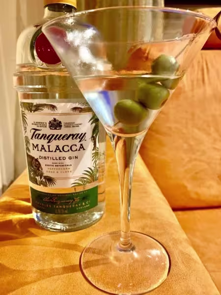A bottle of Tanqueray Malacca gin stands behind a martini glass filled with clear liquid and three green olives, set on a tan surface.