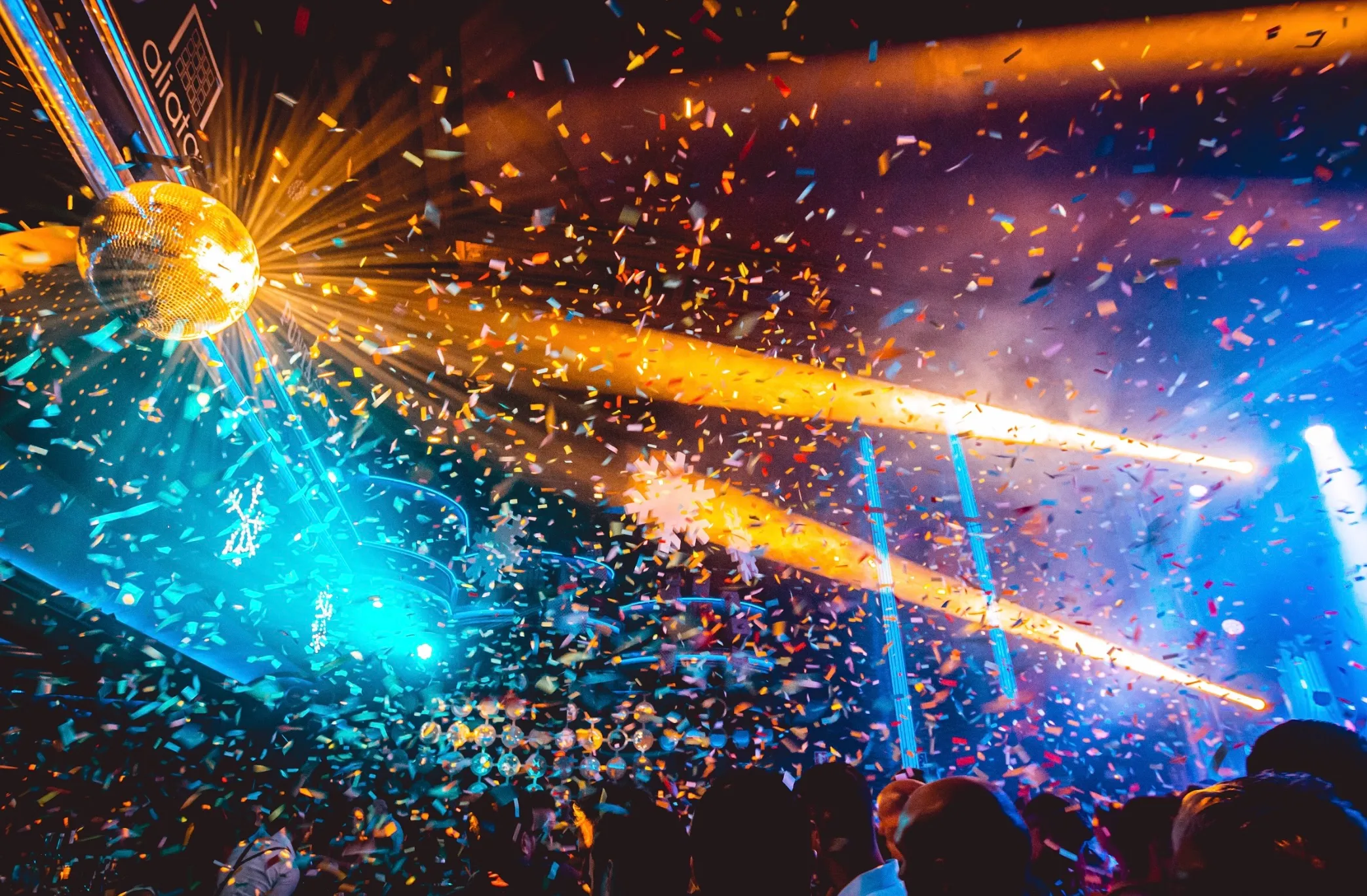 La imagen muestra una discoteca vibrante con una bola de espejos que refleja luces de colores en el ambiente. Confeti cae sobre la multitud mientras haces de luz amarillos y azules crean una atmósfera festiva y llena de energía.