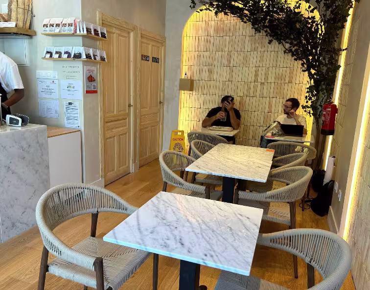 Rincón acogedor de Maison Kayser Padre Damián con sillas de cuerda, mesas de mármol y pared iluminada decorada con vegetación natural.
