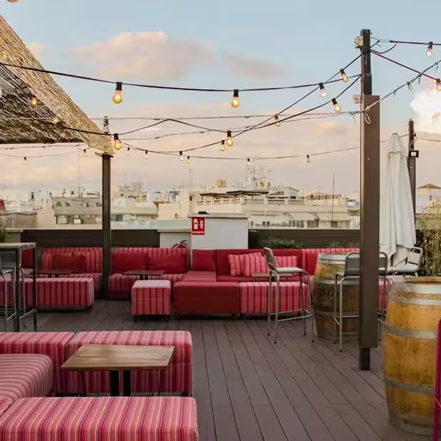 Terraza con sofás a rayas rojas y blancas, barriles como mesas, luces colgantes, sombrillas y vista urbana bajo cielo parcialmente nublado.