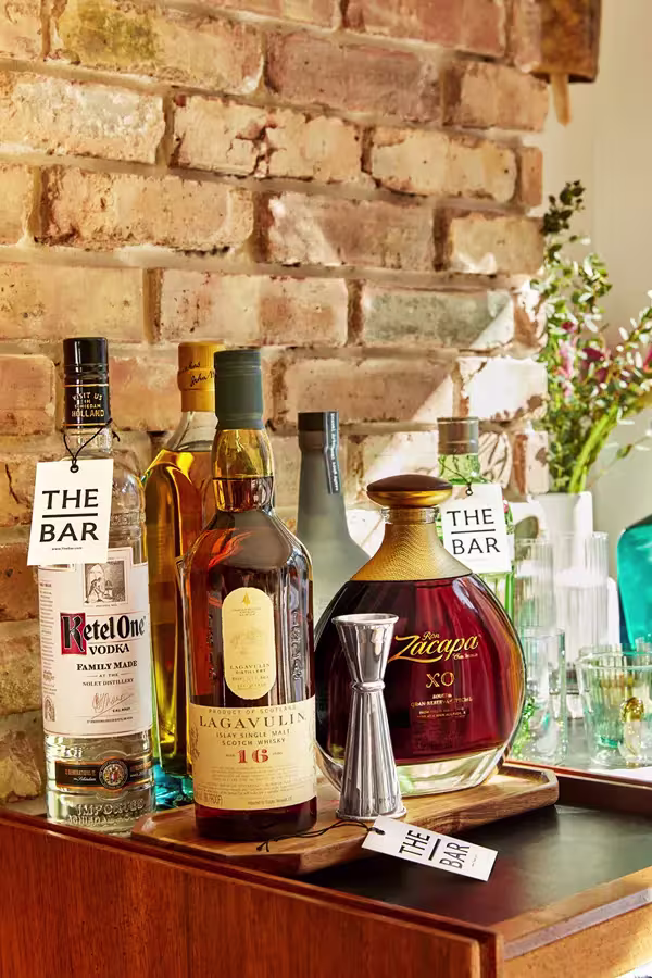 A home bar setup with bottles of Ketel One vodka, Lagavulin 16 whisky, Zacapa XO rum, a jigger, glassware, and The Bar tags, set against an exposed brick wall.