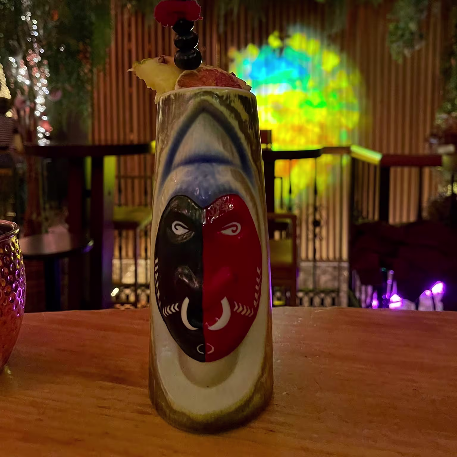 Coctelazo con máscara tribal y frutas frescas bajo la selva luminosa de La Kama Café. Un sorbo, y te transporta.