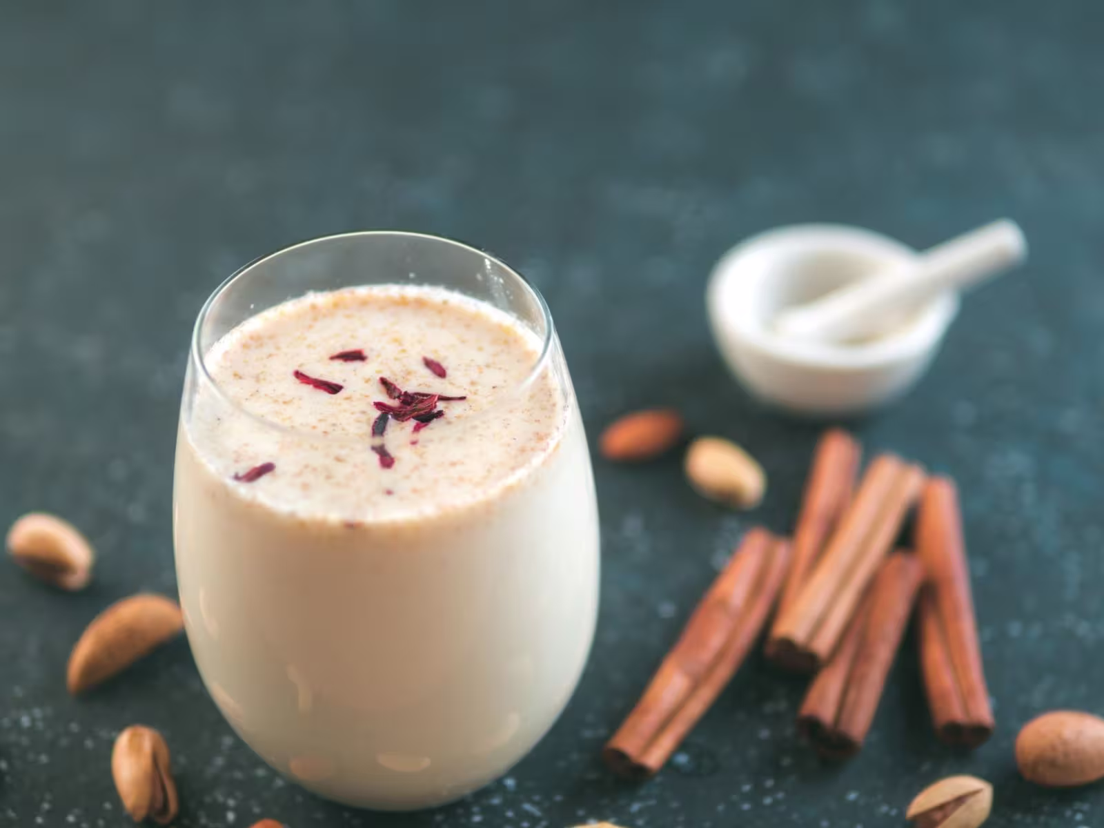 A glass of creamy beverage garnished with saffron strands, placed beside cinnamon sticks, pistachios, and a white mortar and pestle on a dark surface.