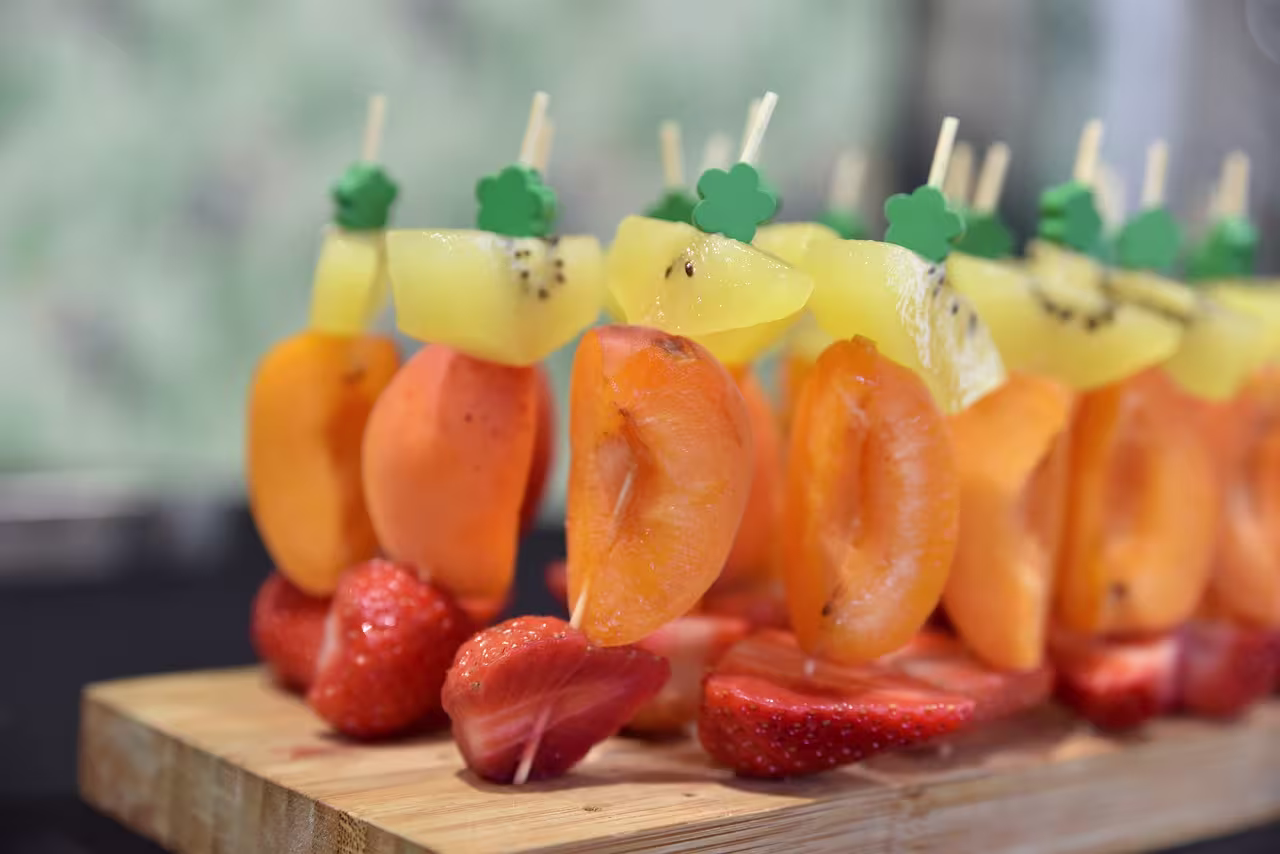 Fruit skewers with slices of apricot, pineapple, and kiwi are arranged on a wooden board, with halved strawberries placed at the base.