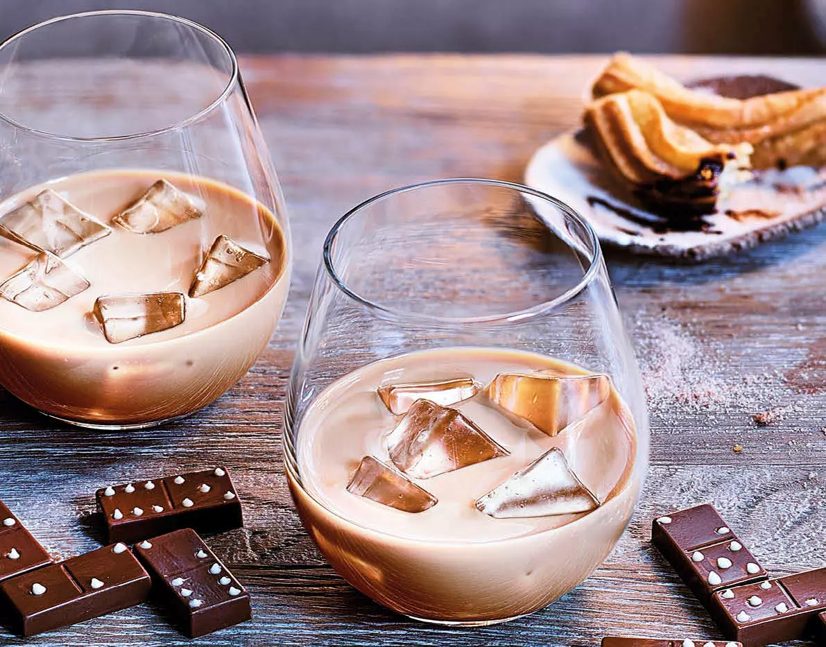 Two glasses of iced creamy liqueur sit on a wooden table with dominoes, chocolate pieces, and a plate of churros in the background.