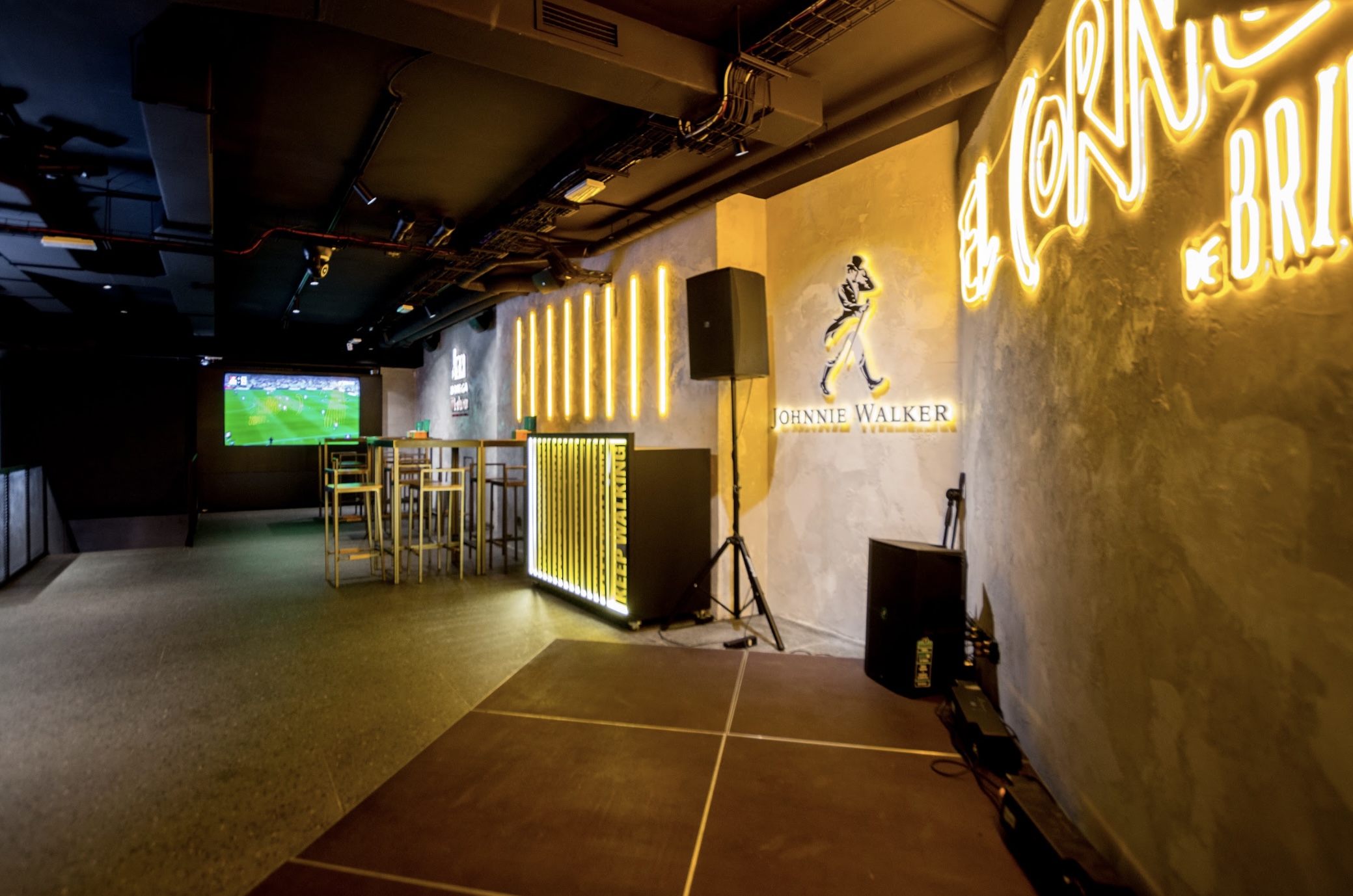 La imagen muestra El Corner del Brindis, un espacio moderno y elegante decorado con neones amarillos y el logo de Johnnie Walker en la pared. Mesas altas y pantallas para ver eventos crean un ambiente ideal para socializar y disfrutar."