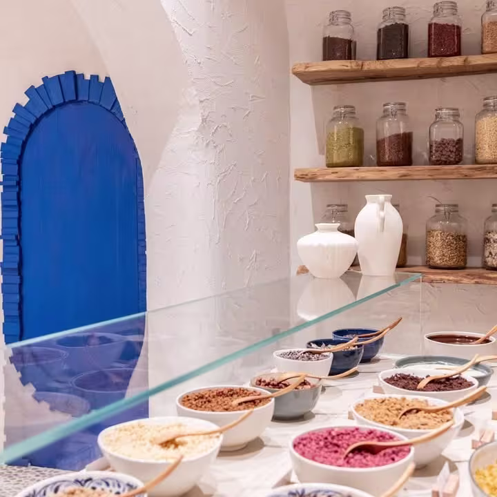 Detalle del mostrador de Myka Ponzano con toppings en cuencos, estanterías rústicas y una puerta azul que contrasta con las paredes blancas.