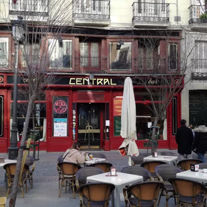 La foto muestra una escena en el interior del Café Central, con cuatro personas sentadas en una mesa. En primer plano, se ve parte una guitarra, lo que sugiere que podría haber música en vivo en el lugar.