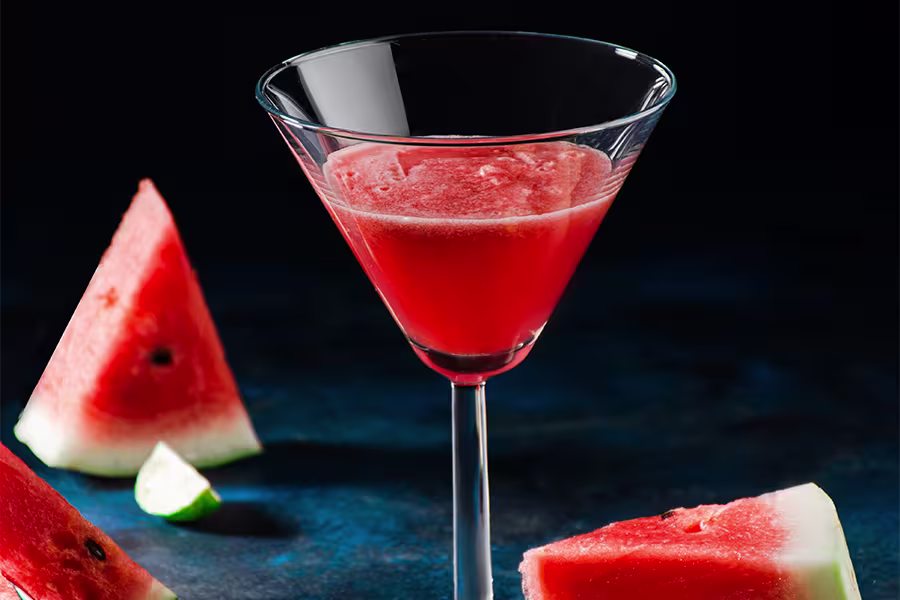 A martini glass filled with a red watermelon cocktail, surrounded by watermelon slices and a lime wedge on a dark surface.