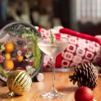 En la imagen se ve un coctel en una copa elegante, alta, con decoración navideña a su alrededor.
