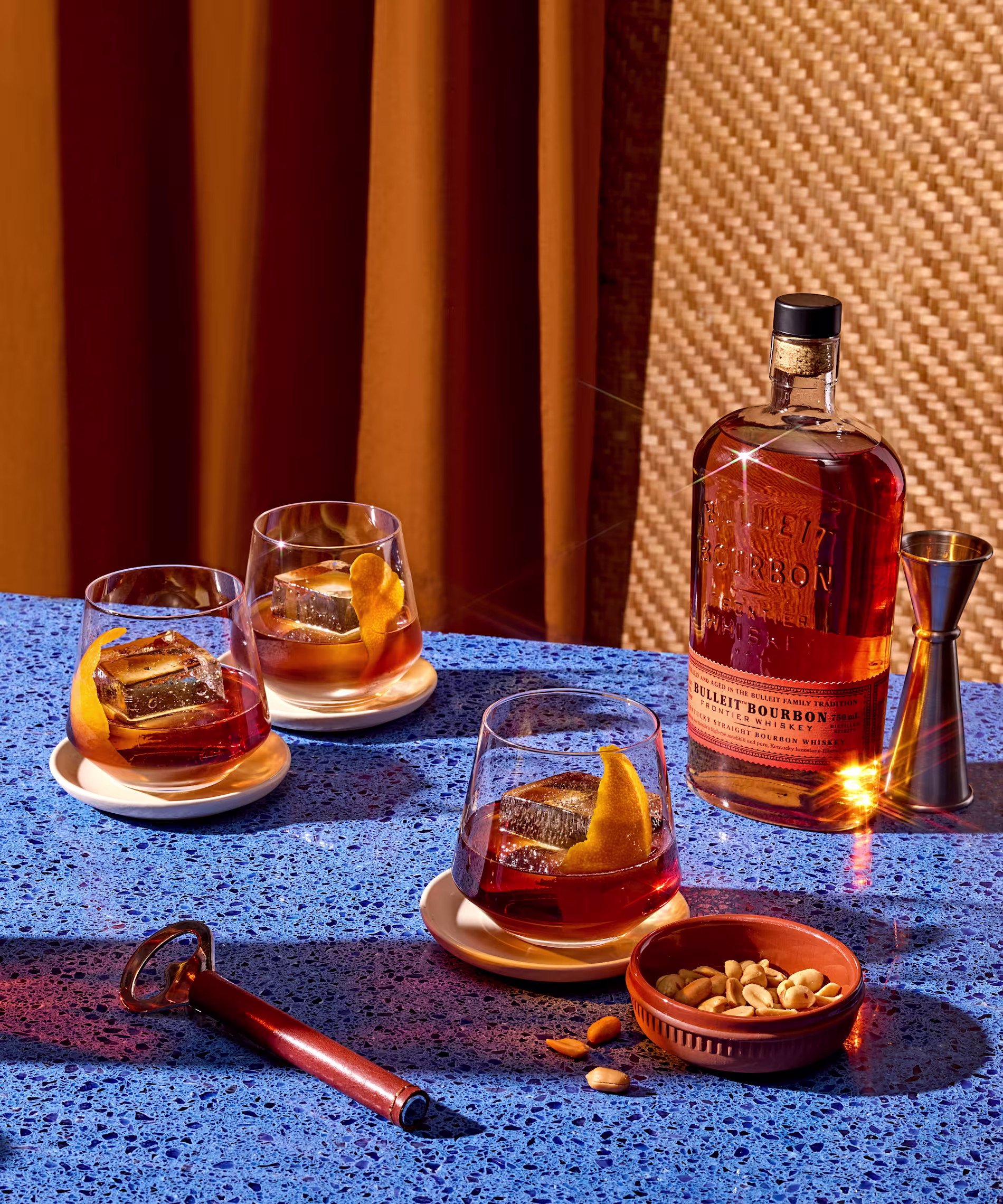 A bottle of Bulleit Bourbon, three glasses with bourbon and orange peel, a jigger, a bowl of mixed nuts, and a cigar cutter on a blue speckled table.