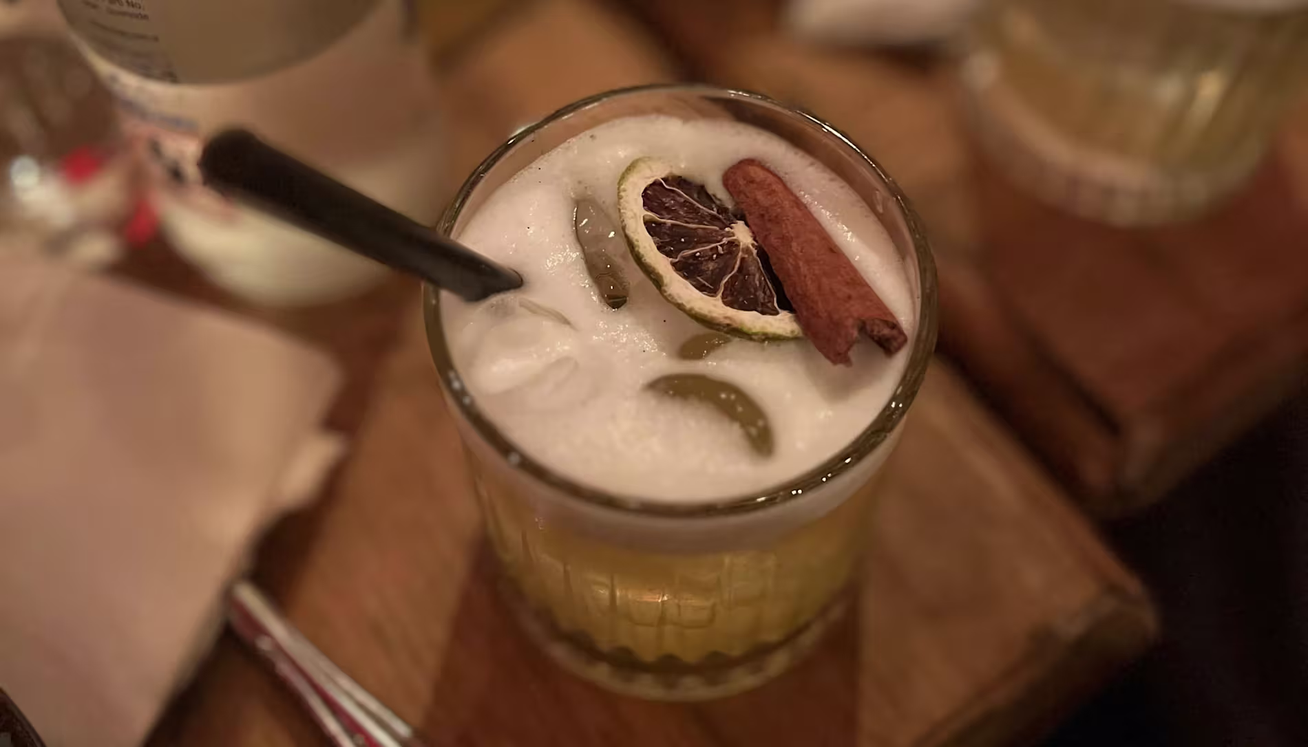 A cocktail with ice, frothy top, dried lime slice, and a cinnamon stick garnish, served in a short glass with a black straw on a wooden surface.