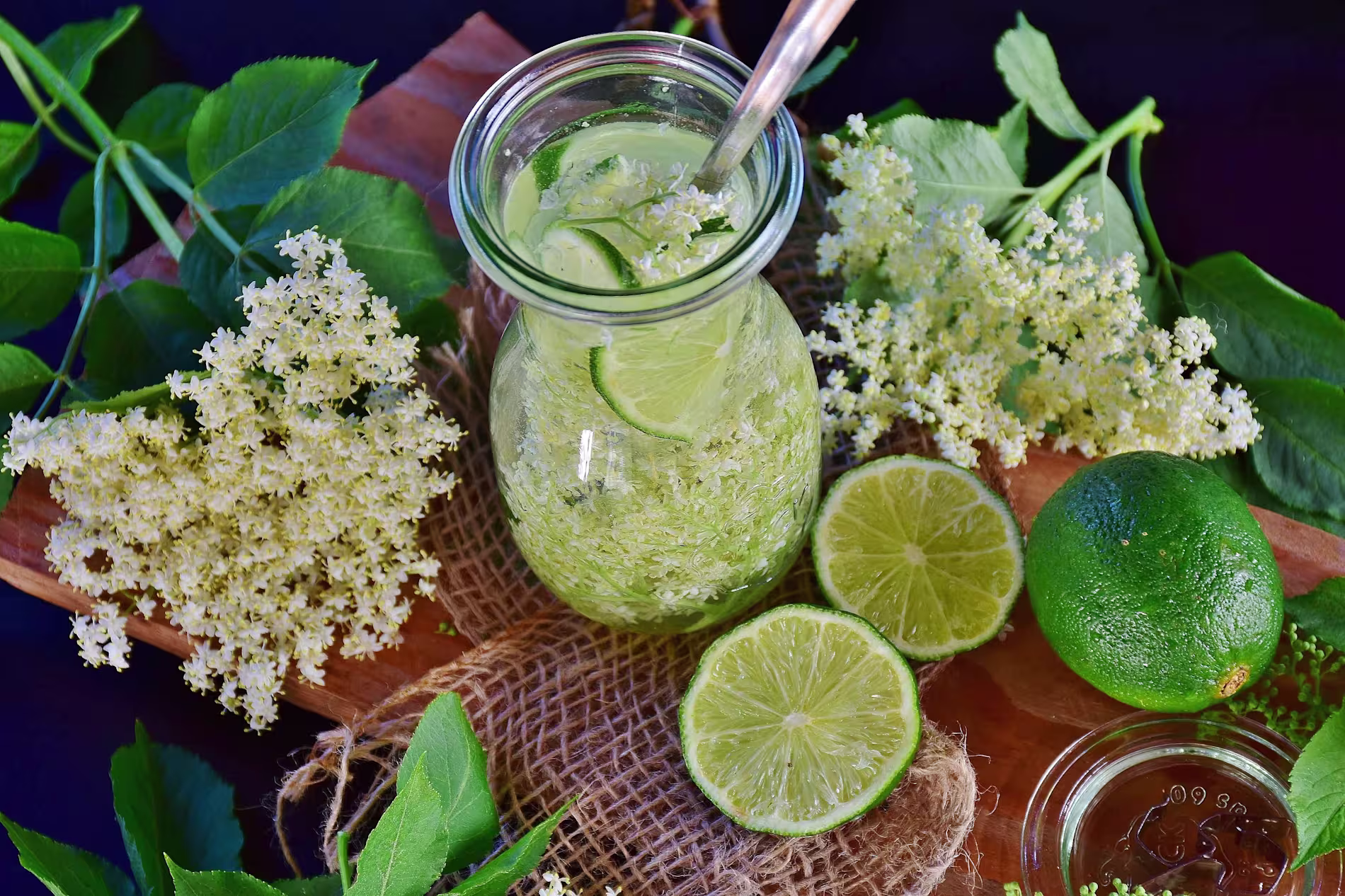 A glass jar with a lime and elderflower drink, surrounded by fresh lime slices, elderflowers, and green leaves on a wooden board.