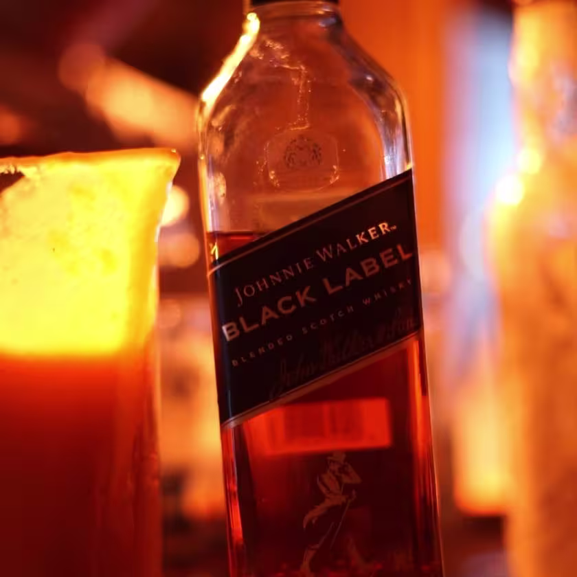 En la imagen se puede observar una botella destapada de Johnnie Walker Black Label junto a un fragmento de una jarra que contiene un cóctel rojizo, sobre una barra de bebidas.