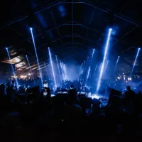 La imagen muestra una vista panorámica de la fiesta con un dj y el público grande enfrente de él.