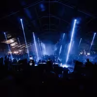 La imagen muestra una vista panorámica de la fiesta con un dj y el público grande enfrente de él.