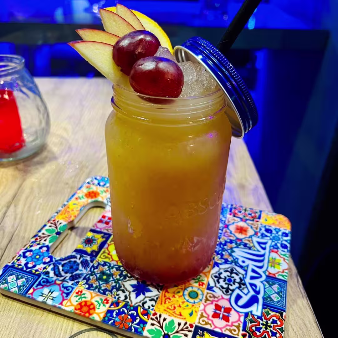 Un cóctel vibrante servido en un tarro, decorado con rodajas de manzana y uvas, sobre una colorida bandeja con motivos de Sevilla.