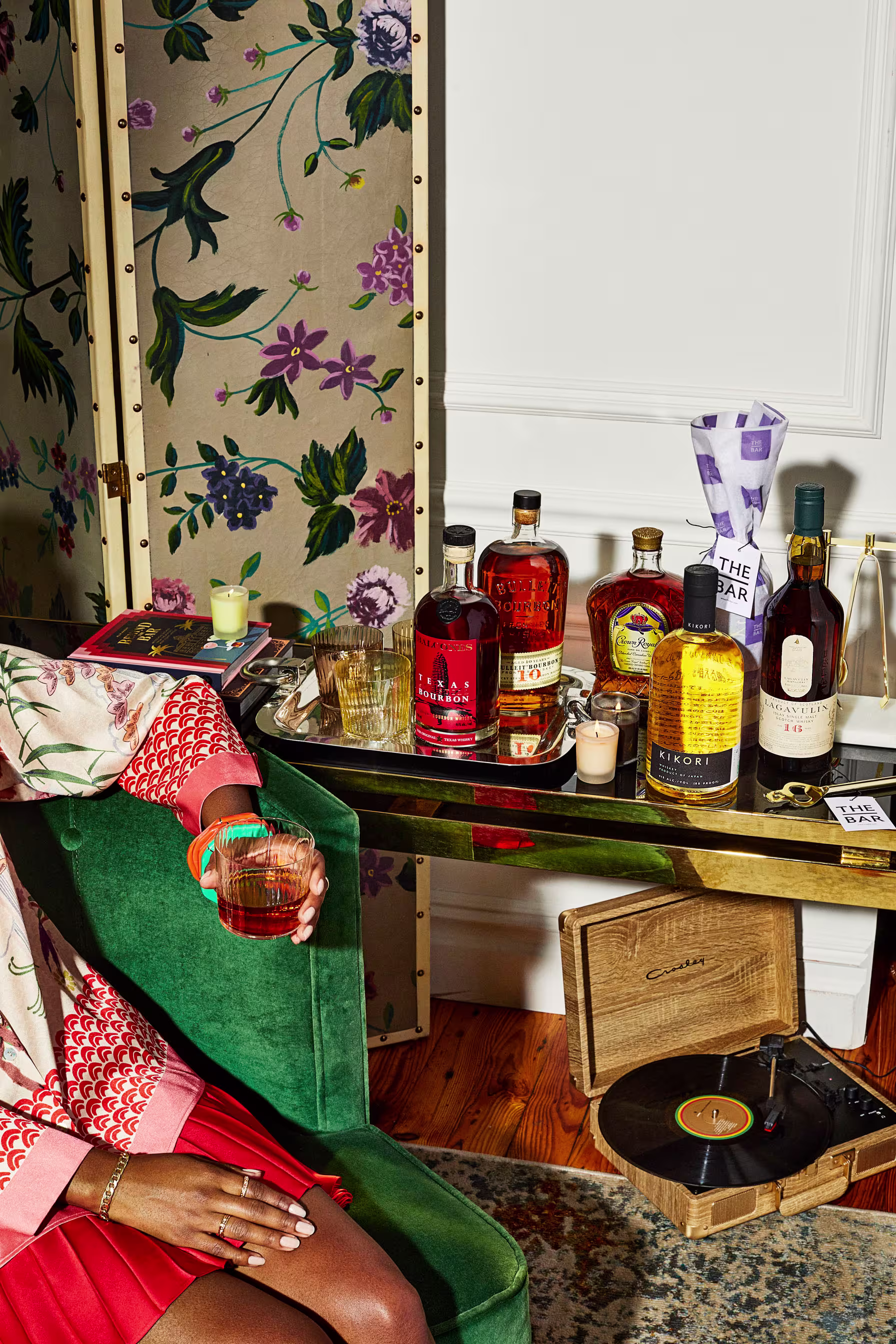 A person holding a drink sits by a table with various liquor bottles, a floral screen, and a record player with a vinyl record on it.
