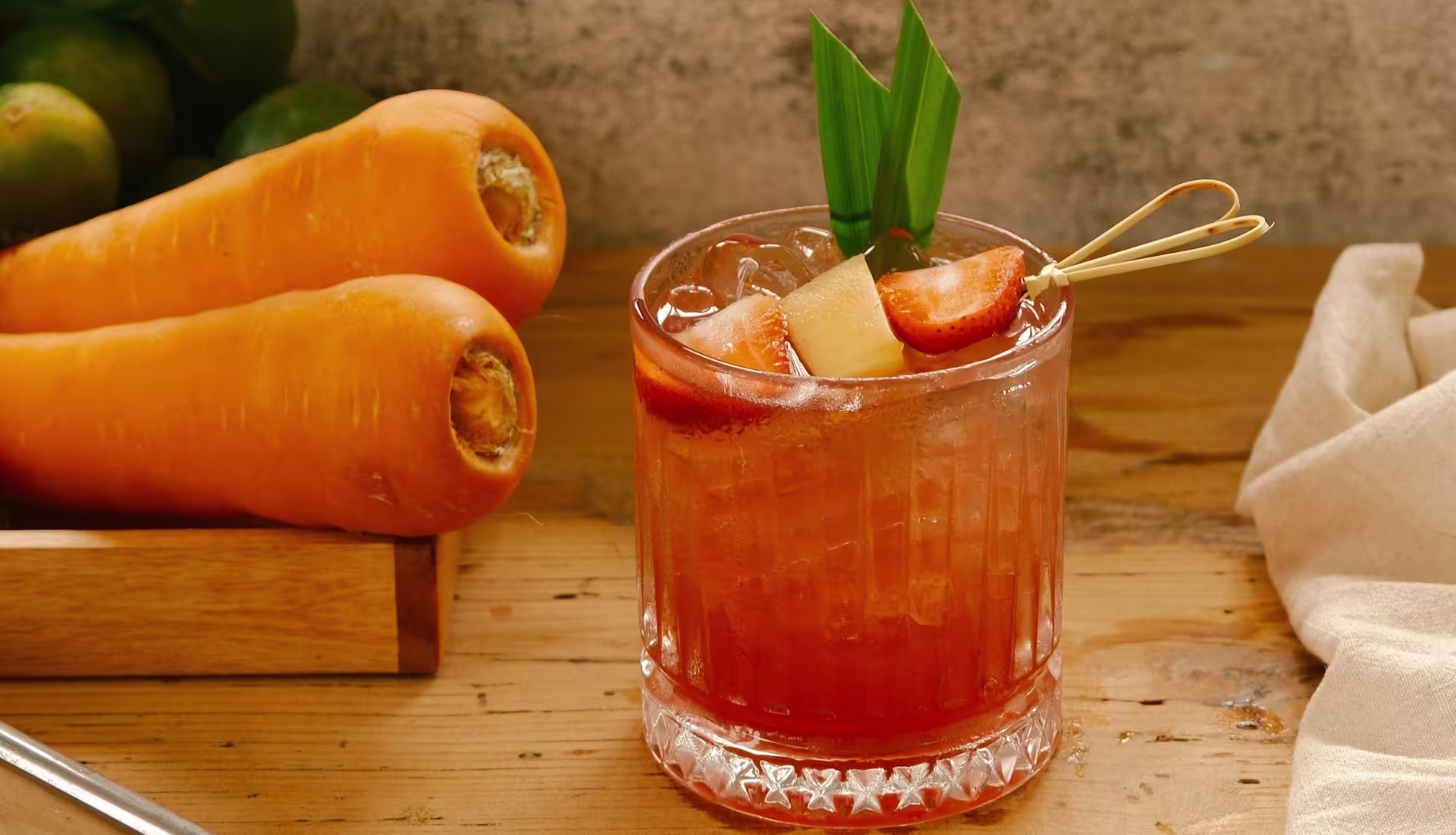 A glass of iced red-orange drink garnished with pineapple, strawberries, and a green leaf, placed on a wooden table next to whole carrots.