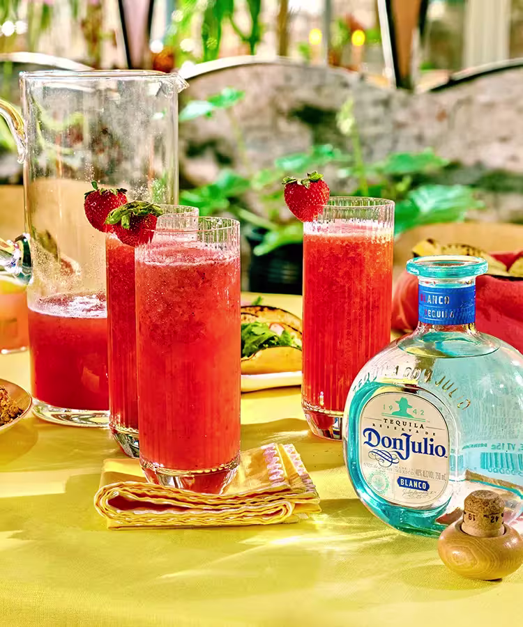 Three glasses of red strawberry cocktails with whole strawberries on the rim, a pitcher, and a bottle of Don Julio Blanco tequila on a yellow tablecloth.