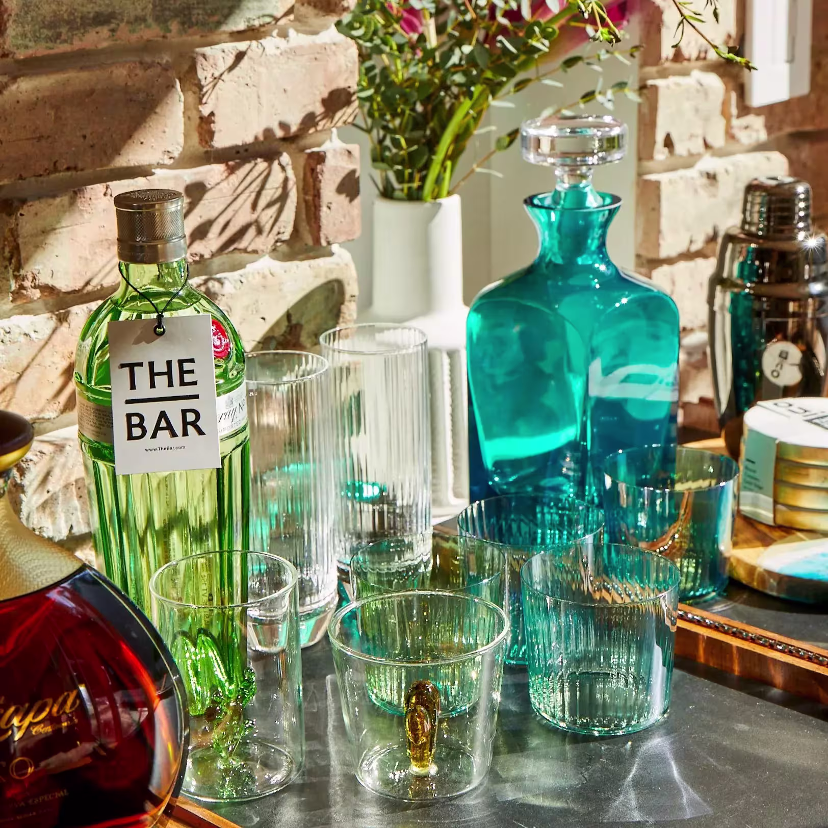 A collection of glassware, a green bottle labeled The Bar, a blue decanter, and cocktail accessories arranged on a table against a brick wall.