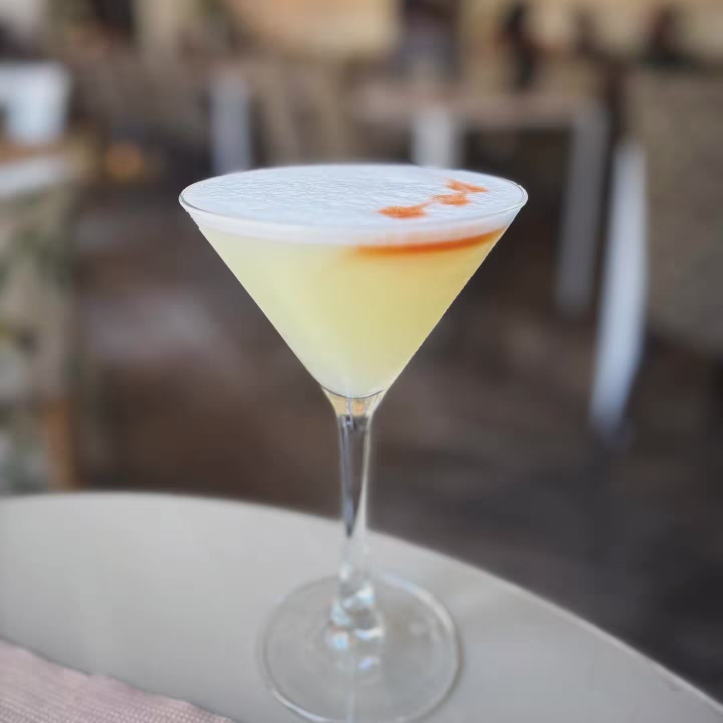 La imagen muestra un primer plano de un cóctel en una copa de martini, que está colocada sobre una superficie con un fondo desenfocado en el interior de El Imparcial.