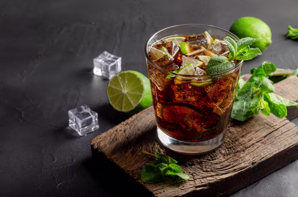 A glass of dark soda with ice, lime slices, and mint sits on a wooden board, with lime, mint, and ice cubes nearby on a dark background.