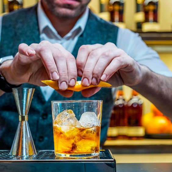 La imagen muestra un bartender en una barra preparando el cóctel Old-Fashioned.