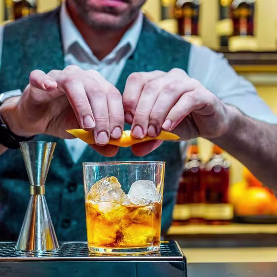 La imagen muestra un bartender en una barra preparando el cóctel Old-Fashioned.