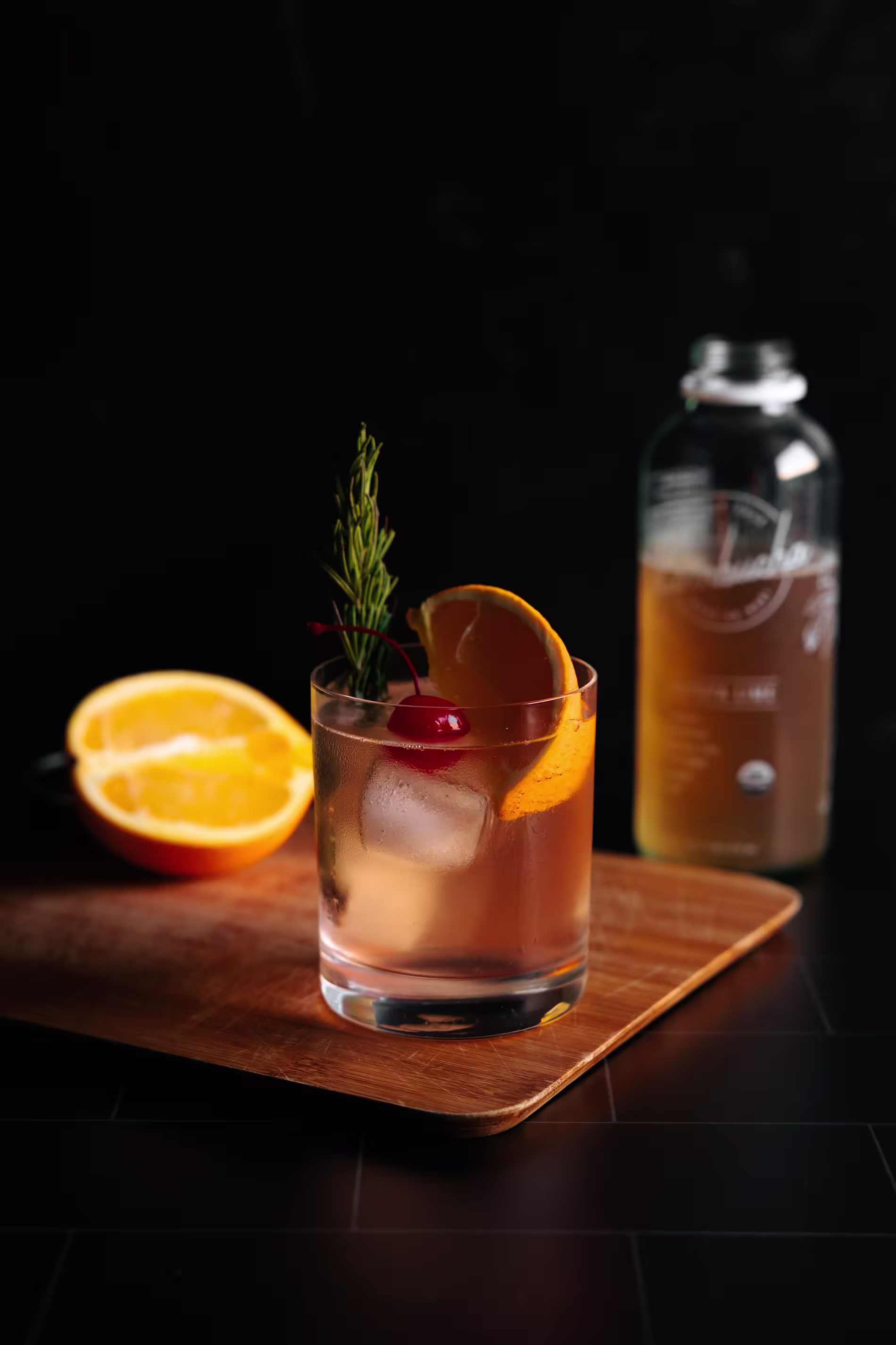 A cocktail with ice, orange slice, cherry, and rosemary in a glass sits on a wooden board, with a halved orange and a beverage bottle in the background.