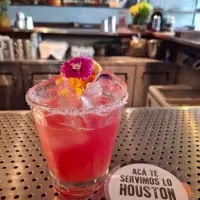 La imagen muestra un colorido coctel sobre la barra decorado con algunas flores y hielo.
