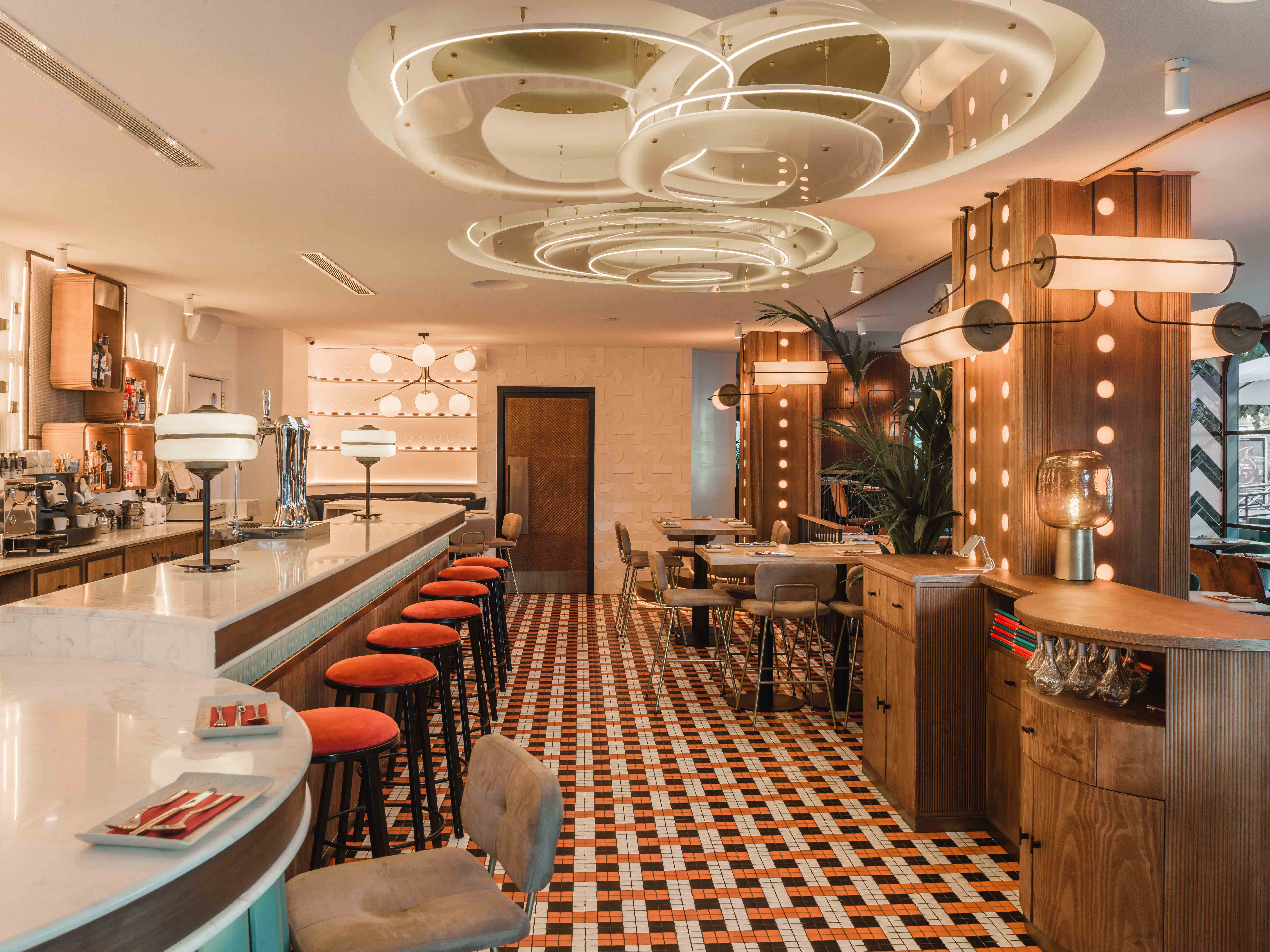 Interior de Ochenta Grados Pozuelo con barra de mármol, taburetes rojos, suelo de mosaico y luminarias circulares que crean un ambiente sofisticado y retro.