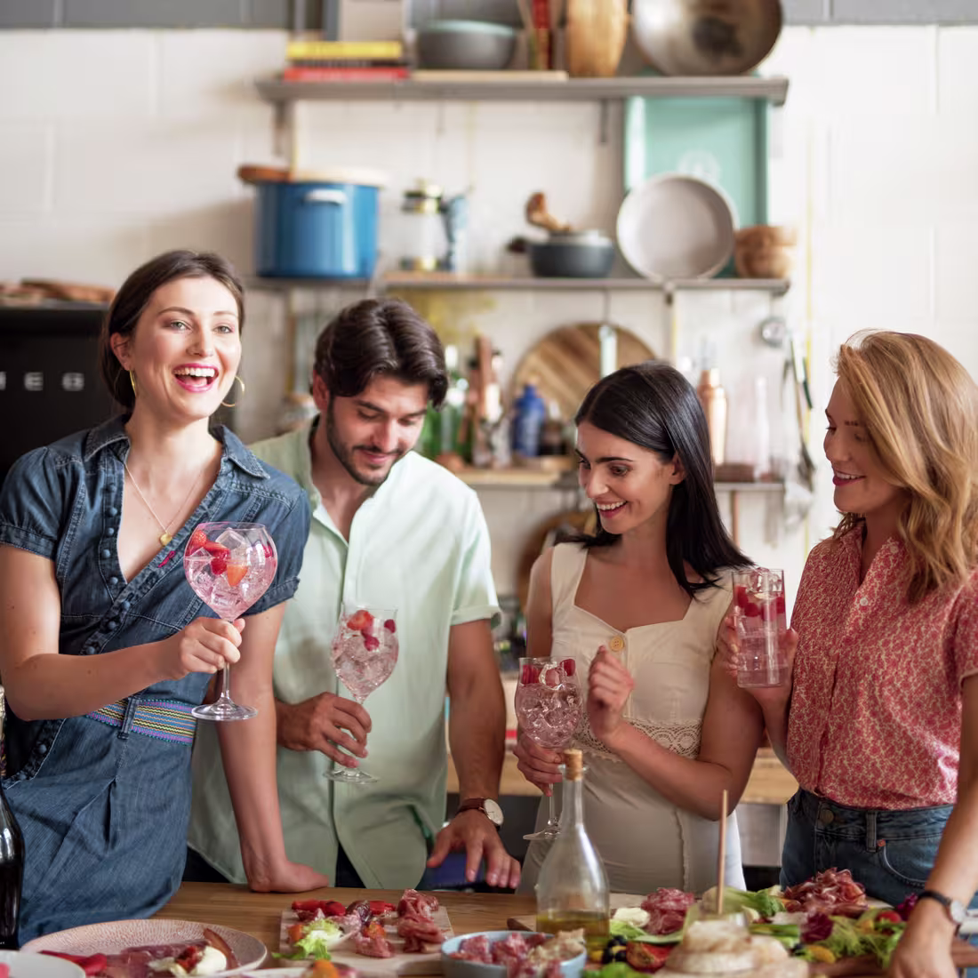 La imagen muestra cuatro personas con distintos cócteles en la mano y una mesa repleta de comida.