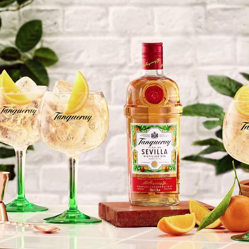La imagen presenta una botella de Tanqueray Flor de Sevilla junto con dos copas que contienen el cóctel Sevilla & Tonic, además de otros elementos de coctelería dispuestos alrededor.