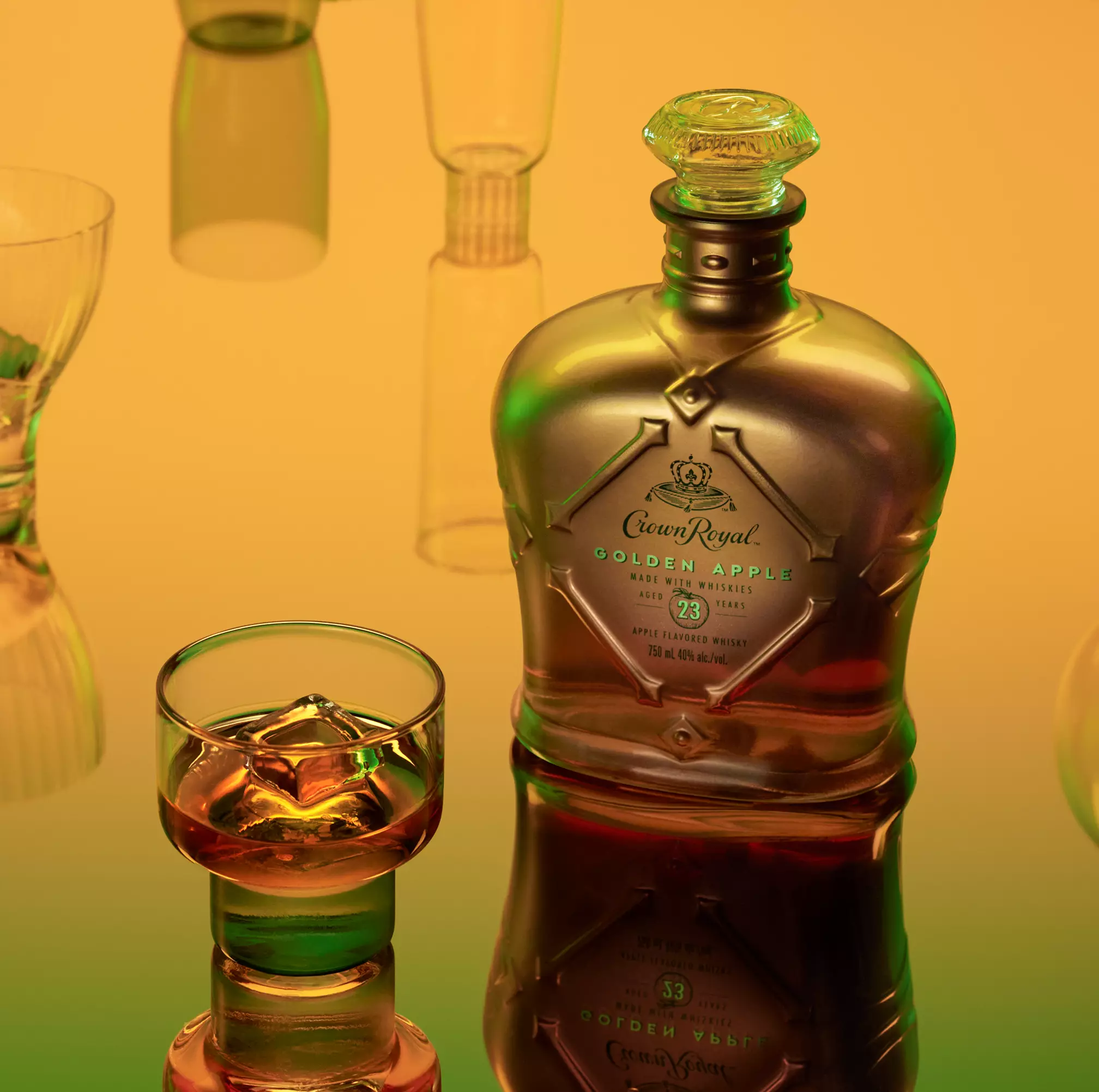 A bottle of Crown Royal Golden Apple whisky and a glass with whisky and ice, set on a reflective surface with a yellow and green gradient background.