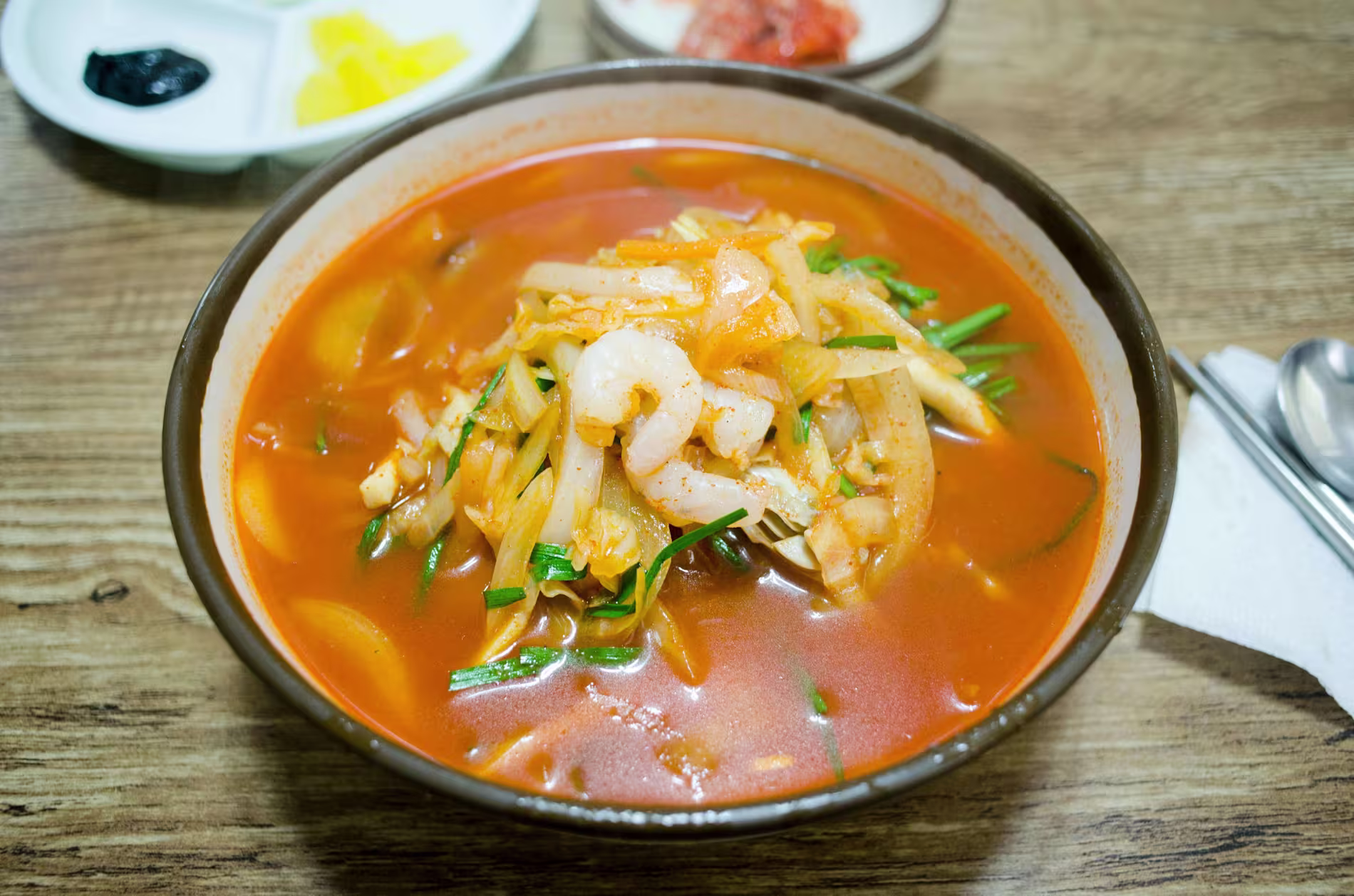 A bowl of spicy red soup with shrimp, vegetables, and noodles on a wooden table, with small side dishes in the background.