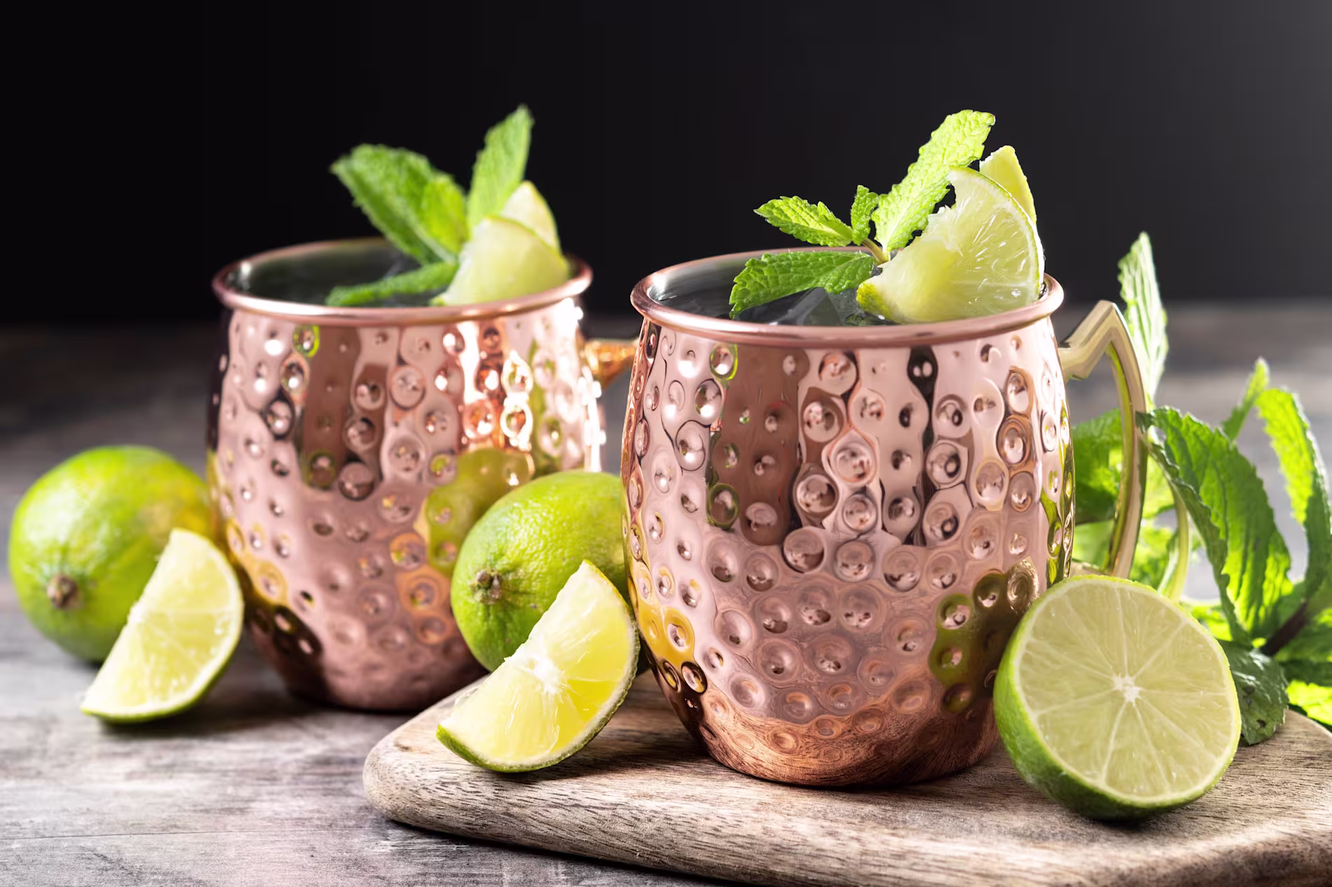 Two copper mugs filled with a drink, garnished with mint and lime, sit on a cutting board surrounded by whole and sliced limes and mint leaves.