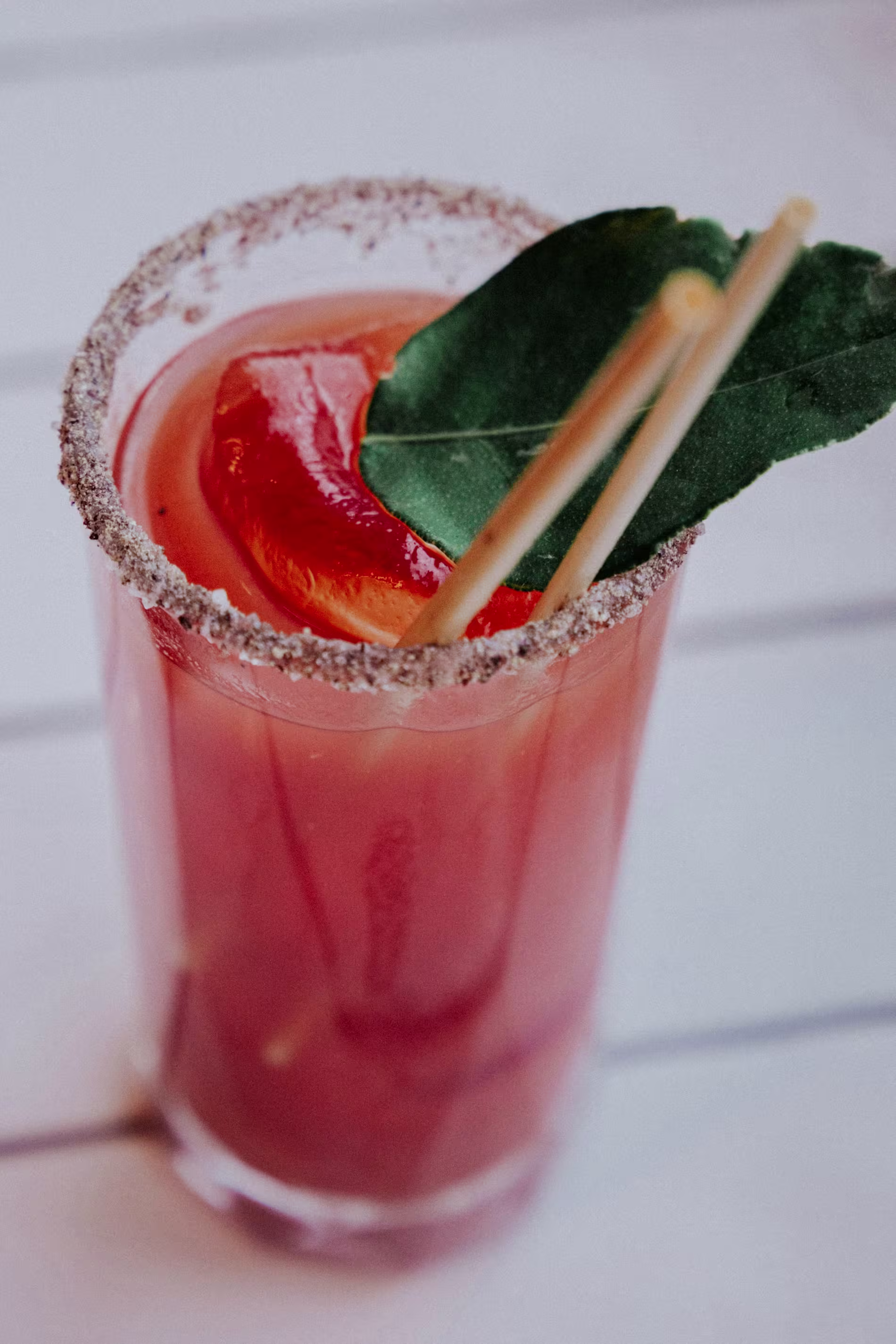 A glass of reddish cocktail with a salted rim, garnished with a citrus slice and a green leaf, and containing two straws.