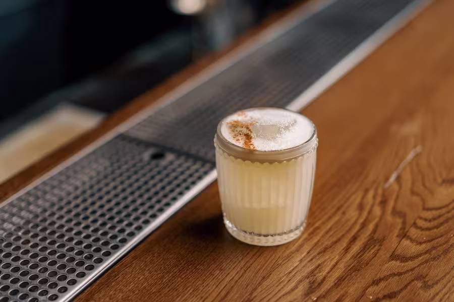 A cocktail with a foamy top and a dusting of spice is served in a patterned glass on a wooden bar counter next to a metal drink rail.