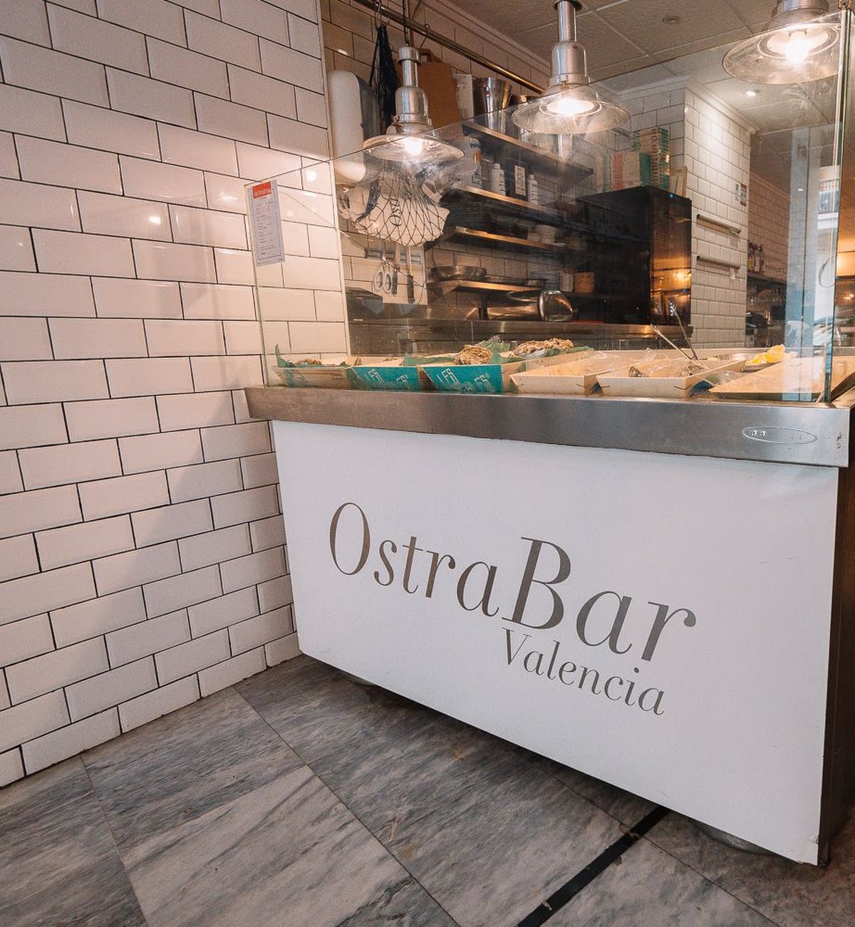 Mostrador de marisco en Ostrabar, con ostras frescas expuestas sobre hielo, baldosas blancas tipo metro y lámparas industriales iluminando la barra.