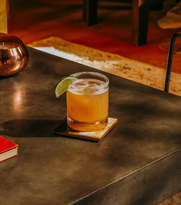 A cocktail garnished with a lime wedge sits on a square coaster on a dark table, with a red book and a candle nearby.