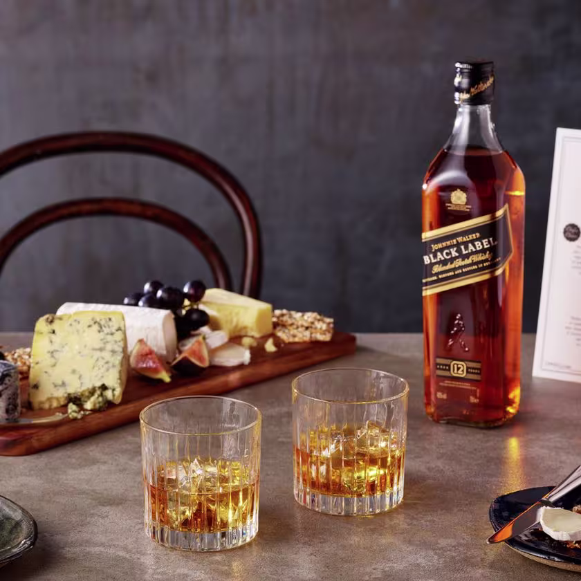 La imagen muestra una tabla de ingredientes salados, una botella de whisky Johnnie Walker Black Label y tres vasos de dicho whisky servido on the rocks.